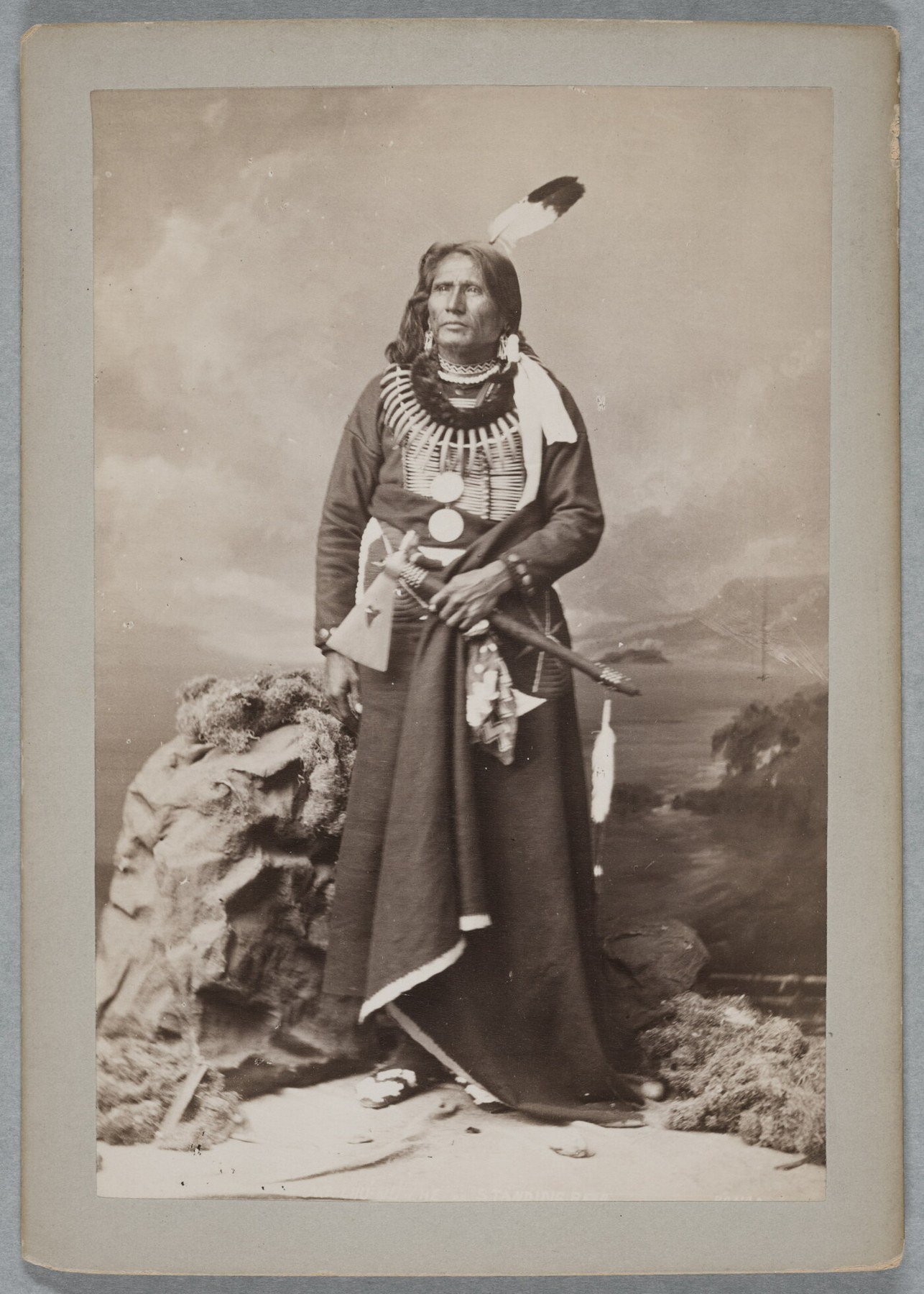 Full-length 1877 studio portrait of Ponca leader Standing Bear wearing a long garment, necklaces and a feather, and holding a pipe tomahawk.